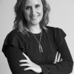 Imagen en blanco y negro de Ángela Gómez Caruncho, directora de Galicia de Torres y Carrera. Tiene el cabello hasta los hombros, vistiendo una camiseta de manga larga y un collar, sonriendo con los brazos cruzados.